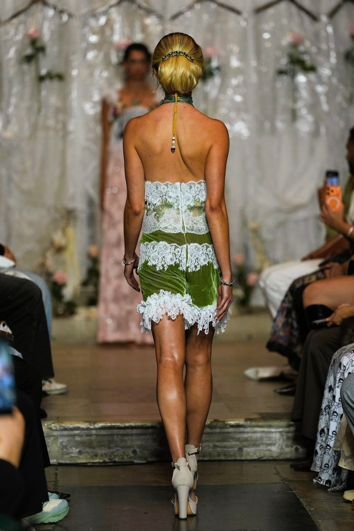 A model viewed from behind walking down a runway wearing a strapless green and white lace mini dress with a halter-style back.