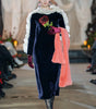 Model walking on a runway wearing a dark blue velvet dress with floral details, white ruffled collar, and pink gloves.