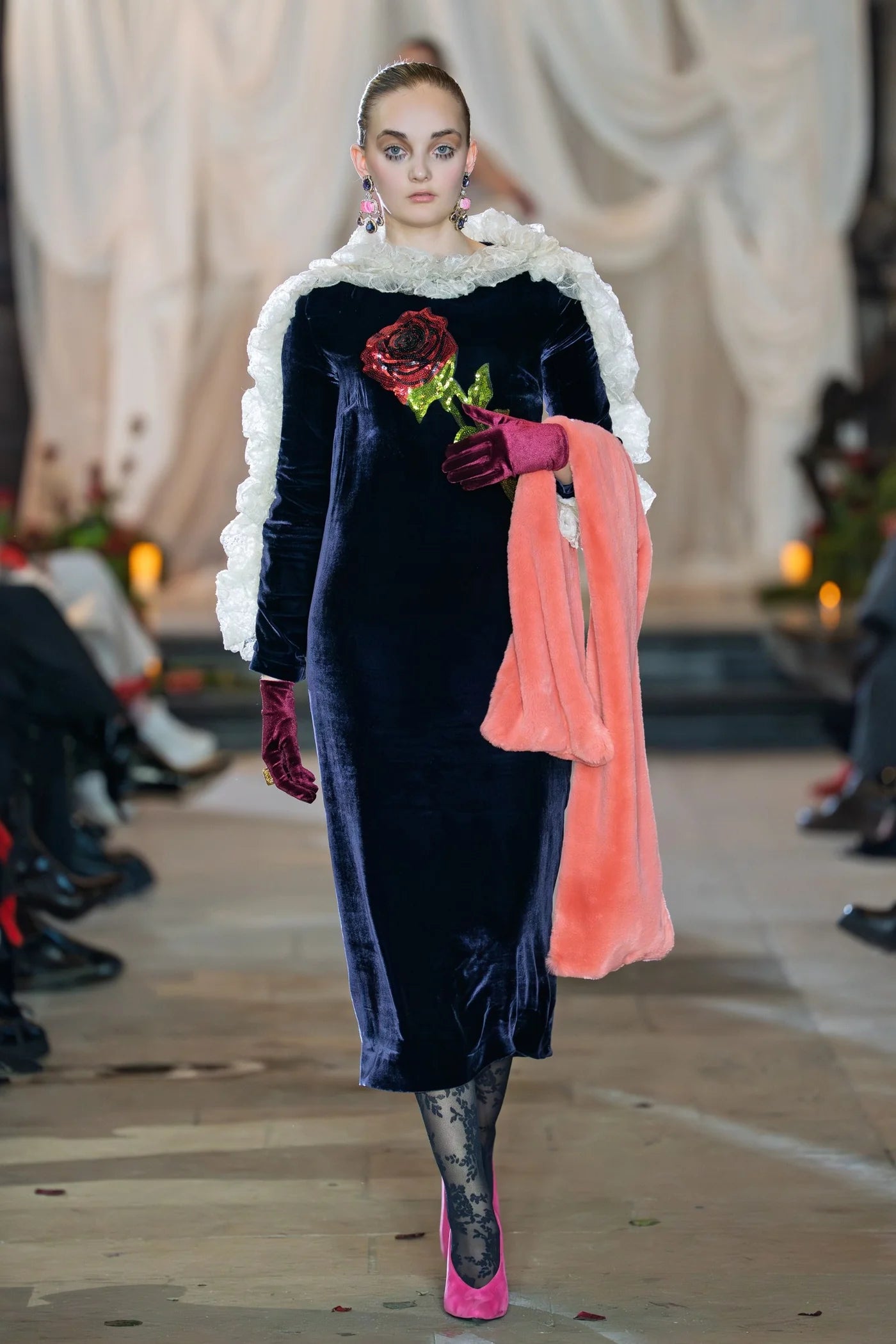 Model walking on a runway wearing a dark blue velvet dress with floral details, white ruffled collar, and pink gloves.