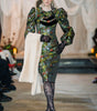 Model wearing a floral dress with long sleeves and black tights on a runway.