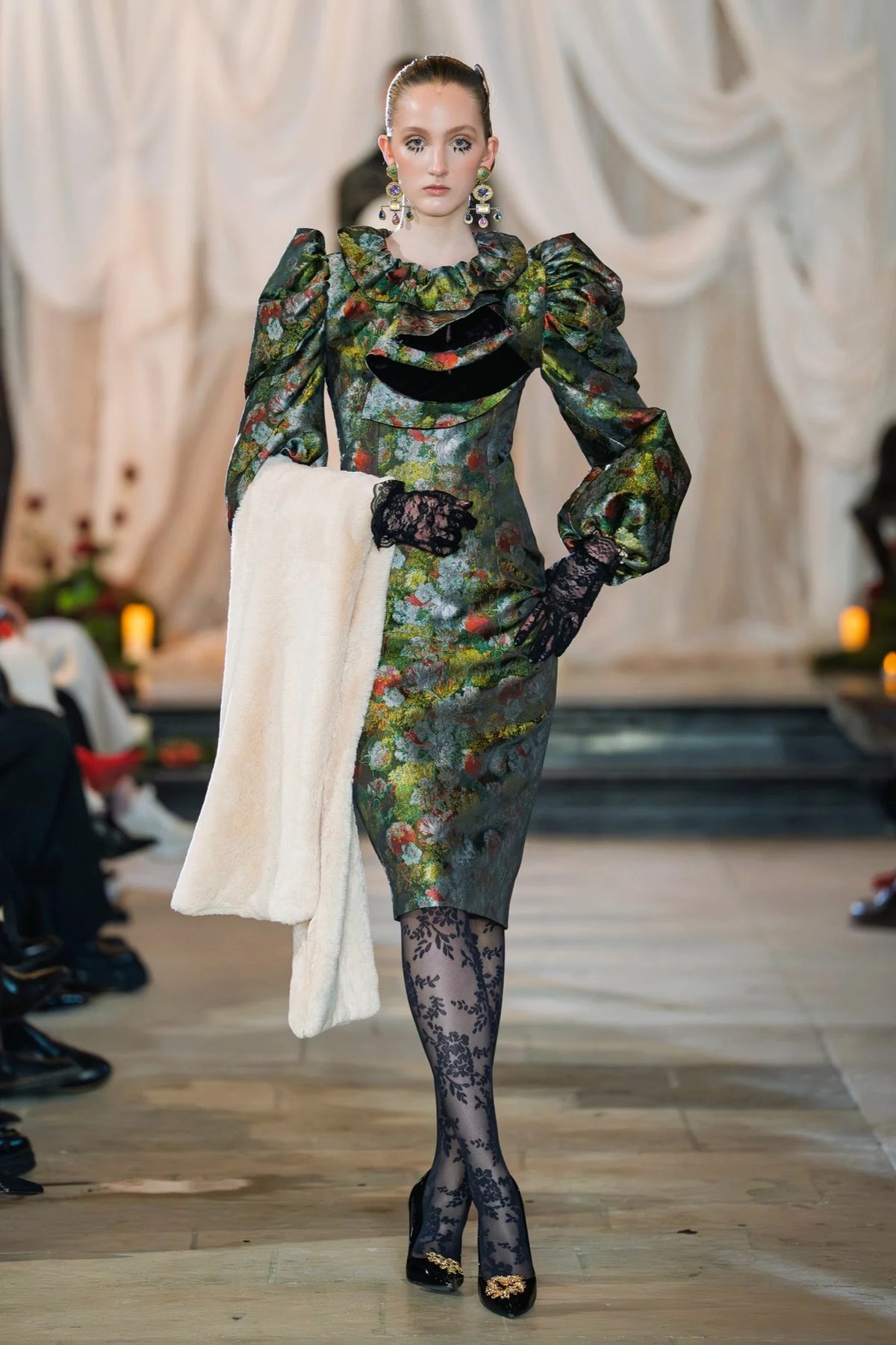 Model wearing a floral dress with long sleeves and black tights on a runway.