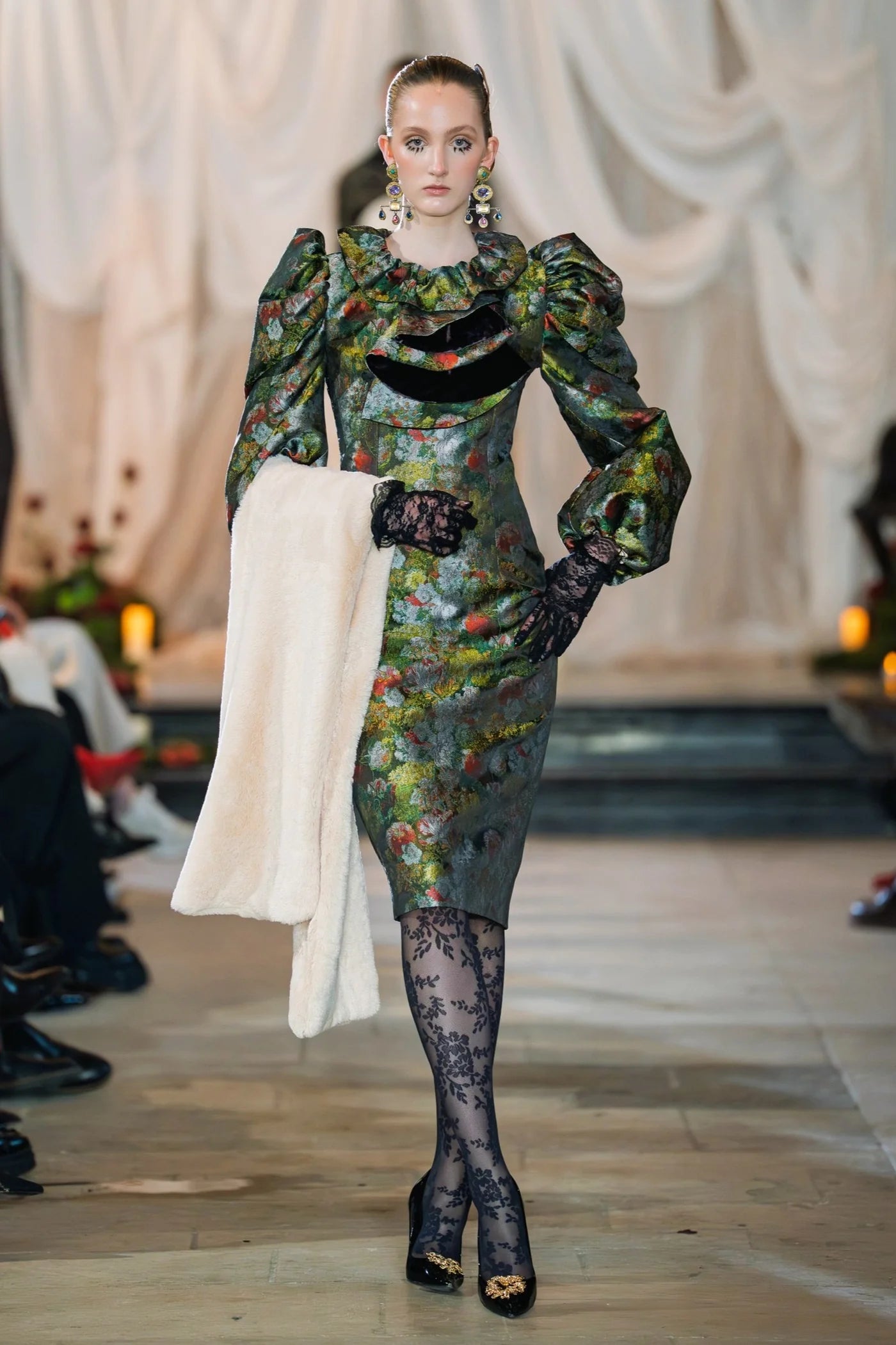 Model wearing a floral dress with long sleeves and black tights on a runway.