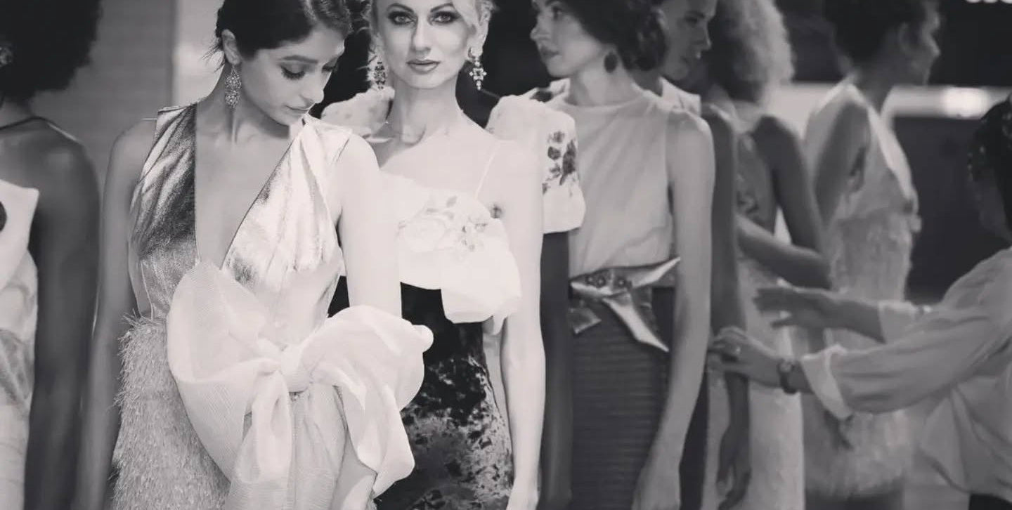 A black and white fashion runway scene with multiple models walking, wearing elegant evening gowns. The central model wears a light-colored dress with a large draped bow detail, surrounded by other fashion shows attendees.