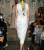 A model viewed from behind walking down a fashion runway wearing a white textured sleeveless dress with green tie details and holding green ribbon streamers. The runway is surrounded by seated spectators holding up cameras.