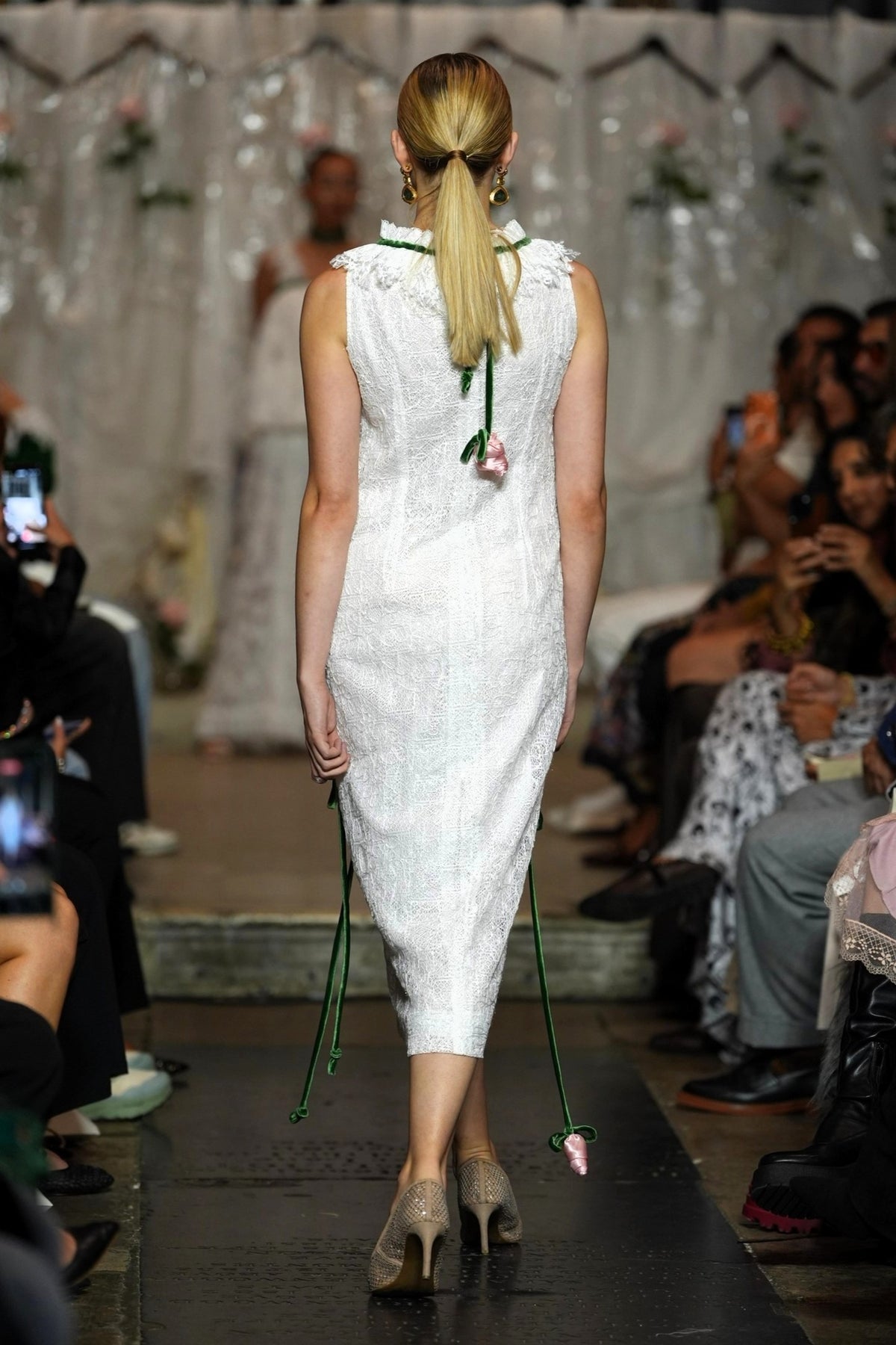 A model viewed from behind walking down a fashion runway wearing a white textured sleeveless dress with green tie details and holding green ribbon streamers. The runway is surrounded by seated spectators holding up cameras.