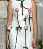 A model walks the runway wearing a sleeveless white textured dress with green trim and pink flower appliqués, with a fashion show audience blurred in the foreground and background.
