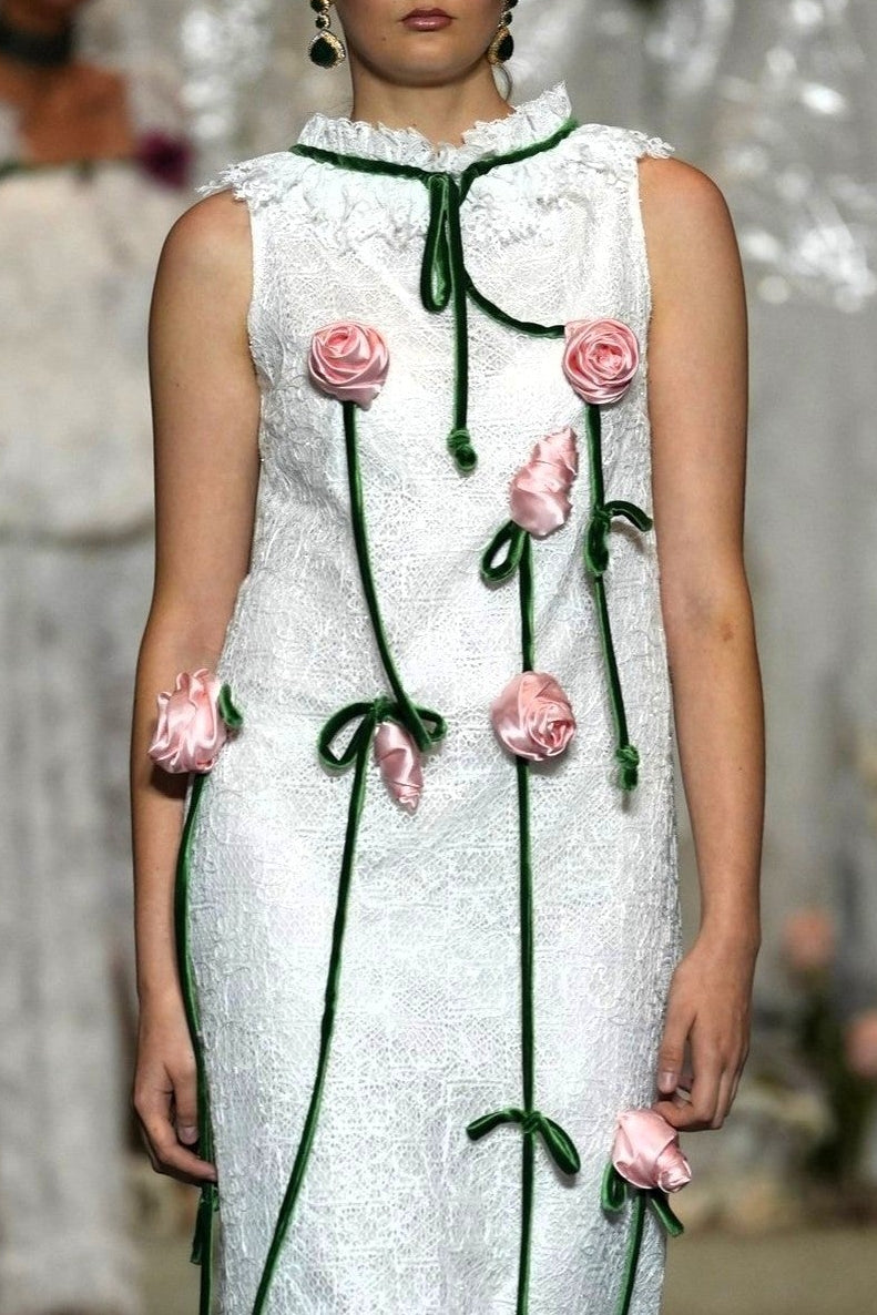 A model walks the runway wearing a sleeveless white textured dress with green trim and pink flower appliqués, with a fashion show audience blurred in the foreground and background.