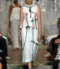 A model walks the runway wearing a sleeveless white textured dress with green trim and pink flower appliqués, with a fashion show audience blurred in the foreground and background.