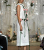 A model walks a fashion runway wearing a sleeveless white textured midi dress decorated with pink roses and green stems draped over the shoulders and sides.