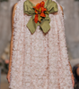 A model walks the runway wearing a sleeveless, textured light pink dress with a large olive green bow and orange floral accent at the neckline, set against a fashion show audience.