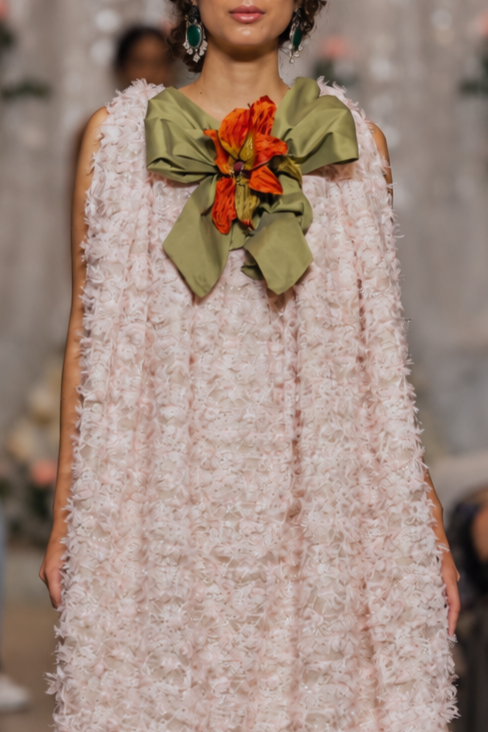 A model walks the runway wearing a sleeveless, textured light pink dress with a large olive green bow and orange floral accent at the neckline, set against a fashion show audience.