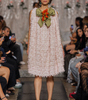 A model walks the runway wearing a sleeveless, textured light pink dress with a large olive green bow and orange floral accent at the neckline, set against a fashion show audience.