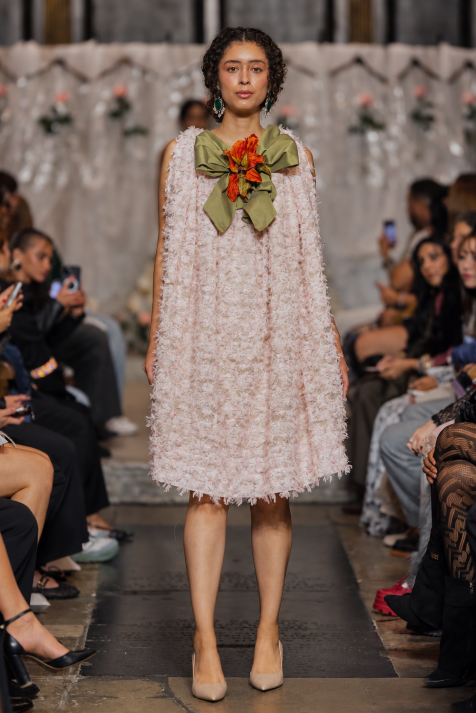 A model walks the runway wearing a sleeveless, textured light pink dress with a large olive green bow and orange floral accent at the neckline, set against a fashion show audience.