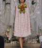 A model walks the runway wearing a light pink textured tulle dress with a large olive green and orange fabric flower accessory pinned at the neckline, set against a runway backdrop with plastic sheeting and floral decorations.
