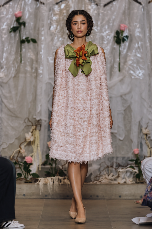 A model walks the runway wearing a light pink textured tulle dress with a large olive green and orange fabric flower accessory pinned at the neckline, set against a runway backdrop with plastic sheeting and floral decorations.