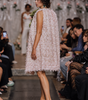A model walks the runway wearing a sleeveless, textured light pink dress with a green bow accent, photographed during a fashion show with seated spectators on both sides.