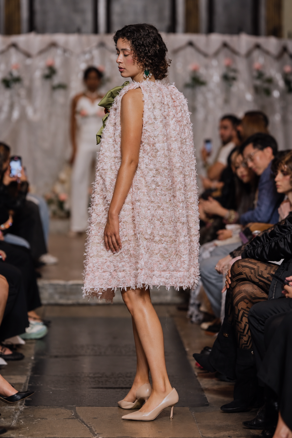 A model walks the runway wearing a sleeveless, textured light pink dress with a green bow accent, photographed during a fashion show with seated spectators on both sides.