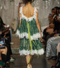 A model viewed from behind wearing a green lace dress with white lace trim and a bright green ribbon tied to her back, walking down a fashion runway with an audience seated on both sides.