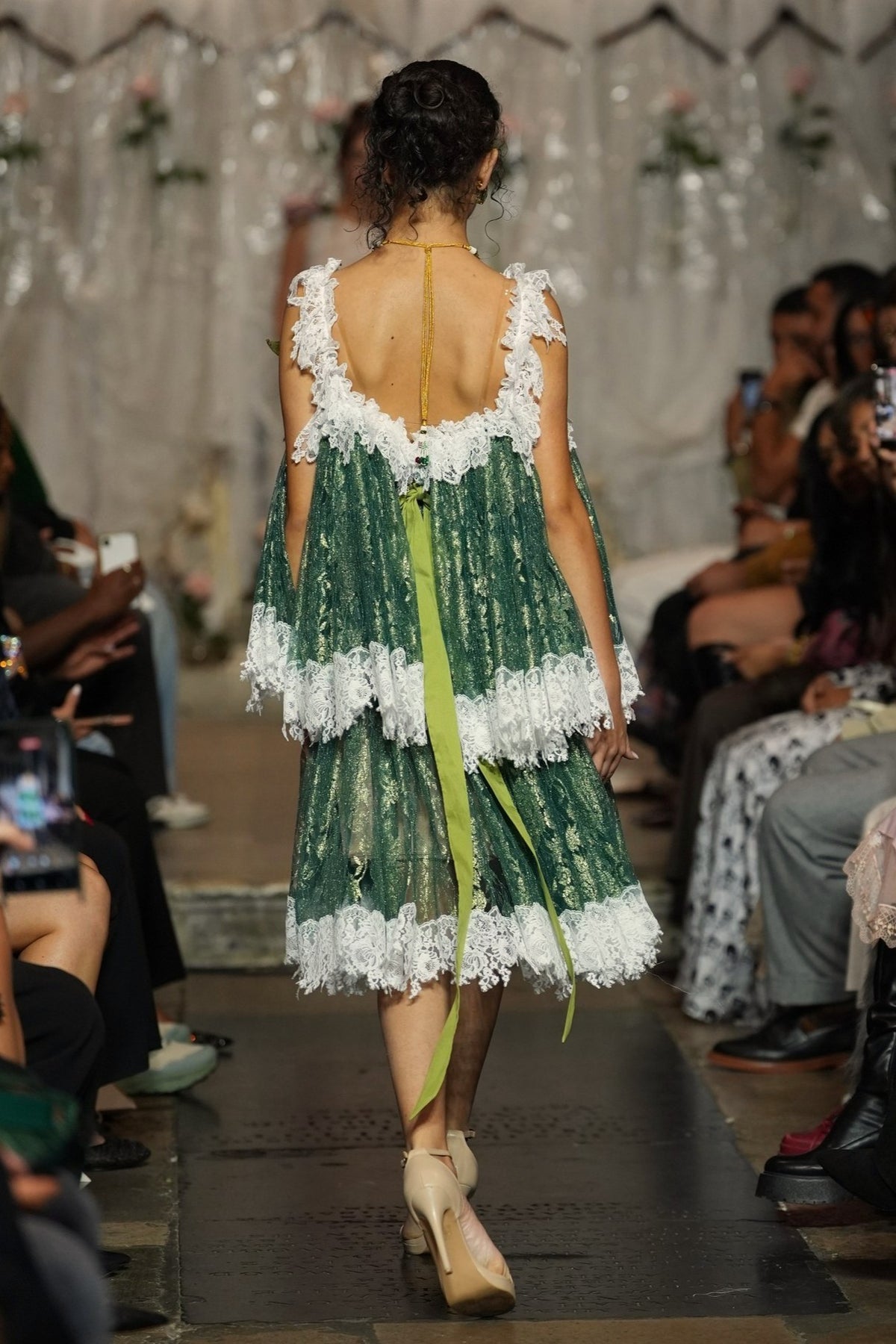 A model viewed from behind wearing a green lace dress with white lace trim and a bright green ribbon tied to her back, walking down a fashion runway with an audience seated on both sides.