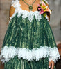 A model walks down a runway wearing a green and white tiered lace dress with off-the-shoulder straps and a floral accessory in one hand. Audience members are seated on both sides of the catwalk.