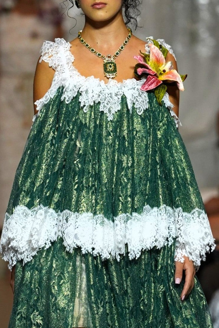 A model walks down a runway wearing a green and white tiered lace dress with off-the-shoulder straps and a floral accessory in one hand. Audience members are seated on both sides of the catwalk.