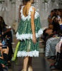 A model from behind wearing a green lace-trimmed dress with a low open back, walking down a fashion runway with an audience seated on both sides.