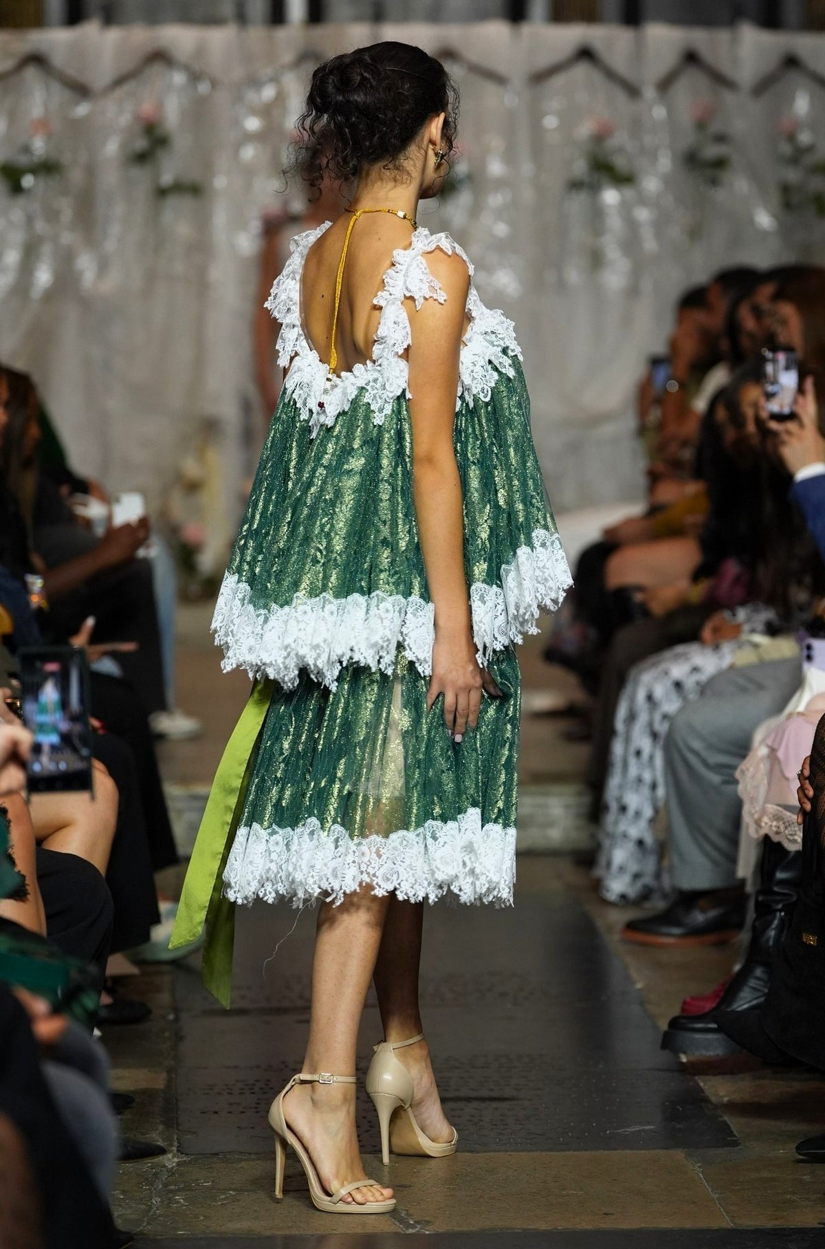 A model from behind wearing a green lace-trimmed dress with a low open back, walking down a fashion runway with an audience seated on both sides.