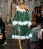 A model walks down a runway wearing a green and white tiered lace dress with off-the-shoulder straps and a floral accessory in one hand. Audience members are seated on both sides of the catwalk.