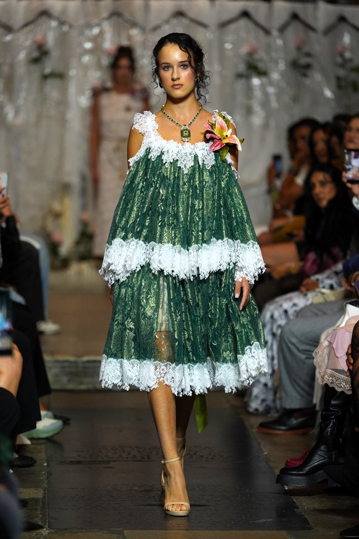 A model walks down a runway wearing a green and white tiered lace dress with off-the-shoulder straps and a floral accessory in one hand. Audience members are seated on both sides of the catwalk.