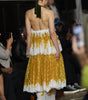 A model from behind wearing a yellow and white tiered lace dress with a green ribbon halter, walking on a fashion runway with audience members in the background.
