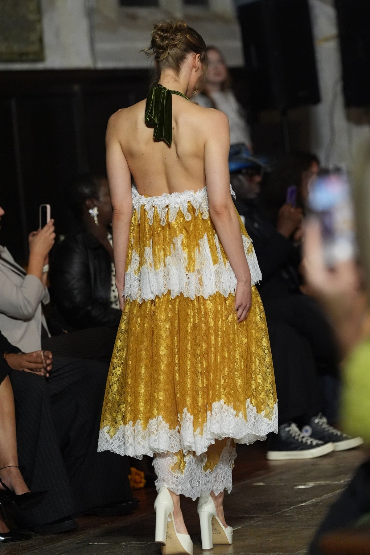 A model from behind wearing a yellow and white tiered lace dress with a green ribbon halter, walking on a fashion runway with audience members in the background.