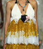 A model walks a fashion runway wearing a halter-neck midi dress with a white lace-trimmed bodice and a voluminous gold skirt with white lace ruffles. The runway show has an audience seated on both sides.