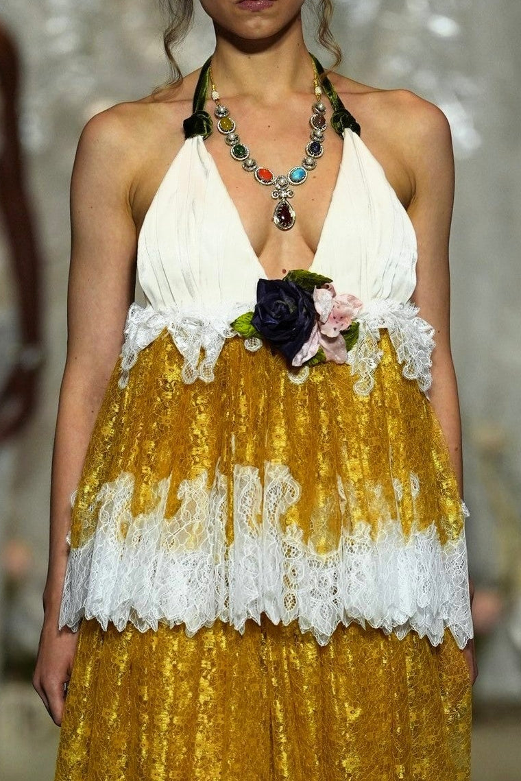 A model walks a fashion runway wearing a halter-neck midi dress with a white lace-trimmed bodice and a voluminous gold skirt with white lace ruffles. The runway show has an audience seated on both sides.
