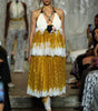 A model walks a fashion runway wearing a halter-neck midi dress with a white lace-trimmed bodice and a voluminous gold skirt with white lace ruffles. The runway show has an audience seated on both sides.
