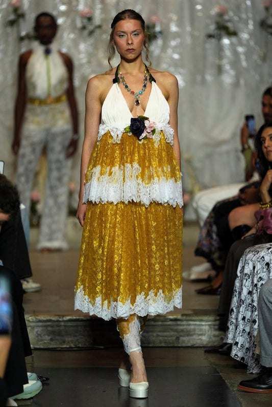 A model walks a fashion runway wearing a halter-neck midi dress with a white lace-trimmed bodice and a voluminous gold skirt with white lace ruffles. The runway show has an audience seated on both sides.