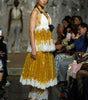 A model walks a fashion runway wearing a sleeveless halter dress with a white upper bodice and layered yellow-gold pleated skirt with white lace trim, paired with white platform heels. Audience members are seated along the runway.