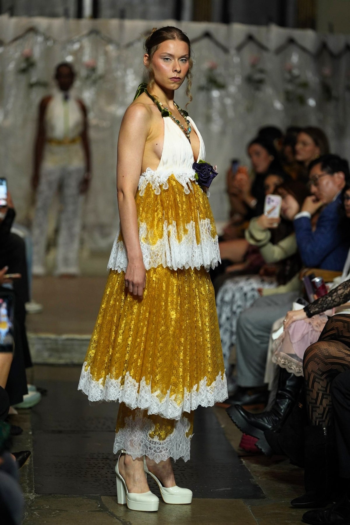 A model walks a fashion runway wearing a sleeveless halter dress with a white upper bodice and layered yellow-gold pleated skirt with white lace trim, paired with white platform heels. Audience members are seated along the runway.