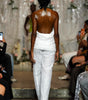 A runway photo showing a model from behind wearing a white strapless top and textured white wide-leg pants, walking on a fashion show catwalk with audience members visible on both sides.