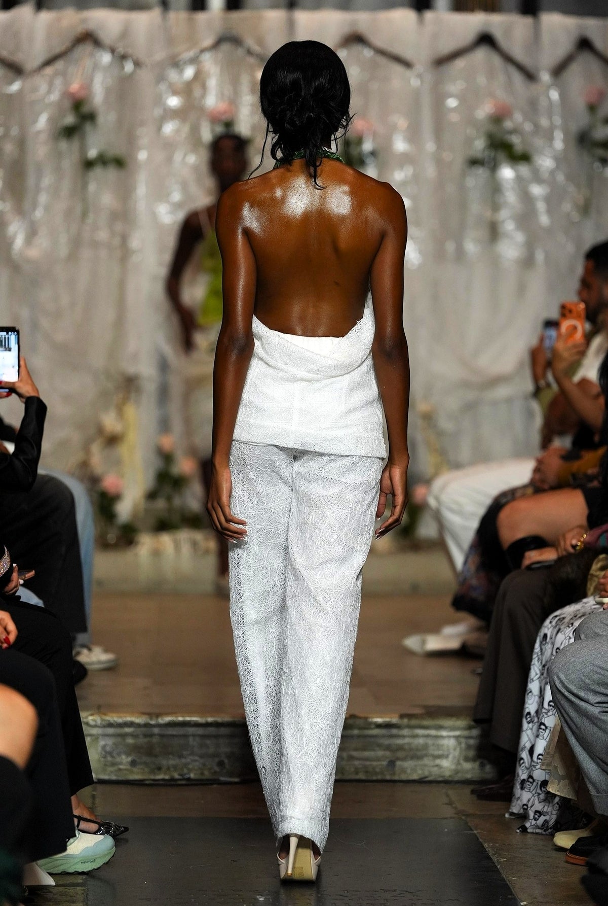 A runway photo showing a model from behind wearing a white strapless top and textured white wide-leg pants, walking on a fashion show catwalk with audience members visible on both sides.