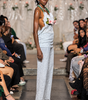 A model walks down a fashion runway wearing a light blue textured sleeveless top and matching wide-leg pants, accessorized with a green beaded necklace and a pink flower in her hair, with seated spectators on both sides.