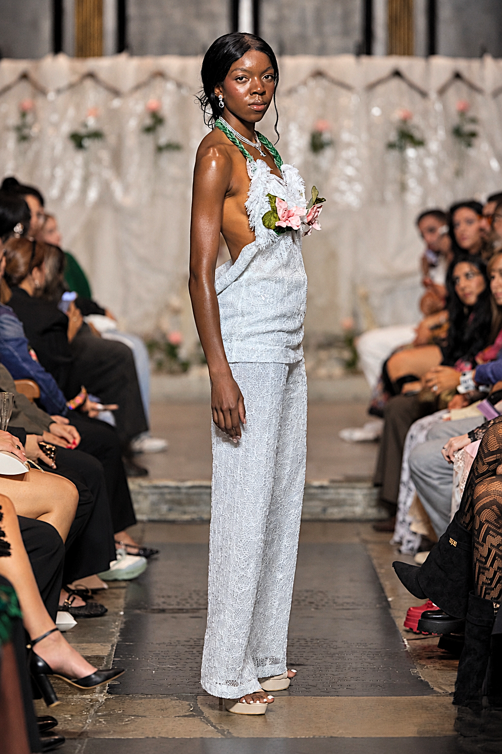 A model walks down a fashion runway wearing a light blue textured sleeveless top and matching wide-leg pants, accessorized with a green beaded necklace and a pink flower in her hair, with seated spectators on both sides.