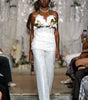 A model walks the runway wearing a white sleeveless top with floral embellishments and matching textured white pants, with seated audience members visible on both sides of the catwalk.