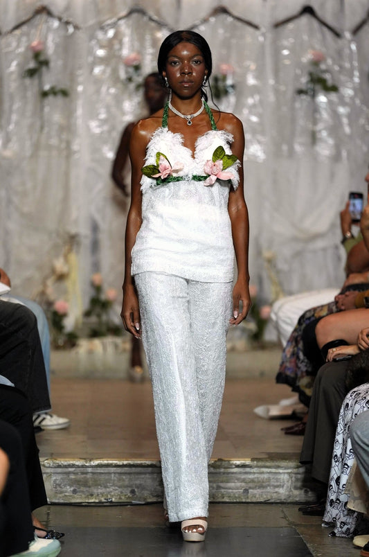 A model walks the runway wearing a white sleeveless top with floral embellishments and matching textured white pants, with seated audience members visible on both sides of the catwalk.