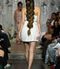 A model viewed from behind on a fashion runway, wearing a strapless white textured mini dress with a very long, thick side-swept braid. The runway is flanked by seated spectators, and another model is visible in the blurred background.