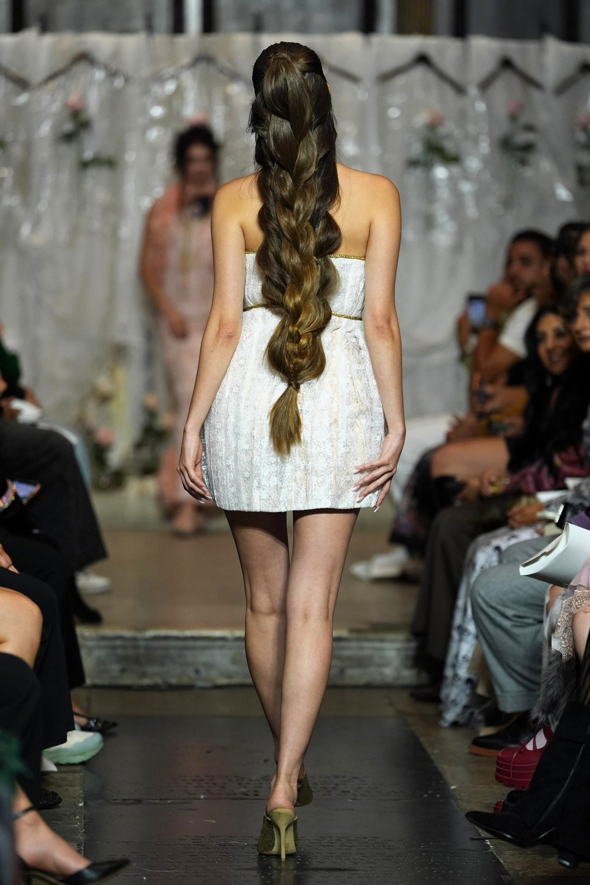 A model viewed from behind on a fashion runway, wearing a strapless white textured mini dress with a very long, thick side-swept braid. The runway is flanked by seated spectators, and another model is visible in the blurred background.
