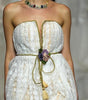 A model walks the runway wearing a strapless white textured mini dress with gold trim and a tassel tie at the waist, photographed during a fashion show with seated spectators in the background.