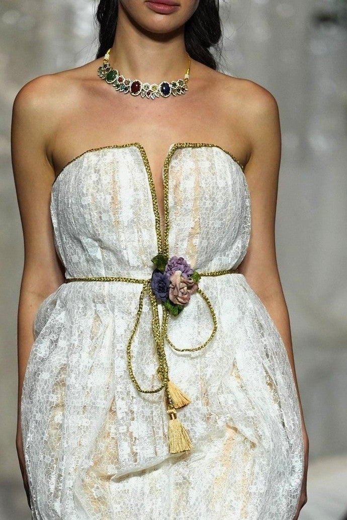 A model walks the runway wearing a strapless white textured mini dress with gold trim and a tassel tie at the waist, photographed during a fashion show with seated spectators in the background.