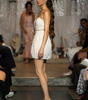 A model walks the runway wearing a strapless white textured mini dress with a gold waist detail and olive green heeled mule sandals. The runway is flanked by seated spectators, with a draped white fabric backdrop in the background.