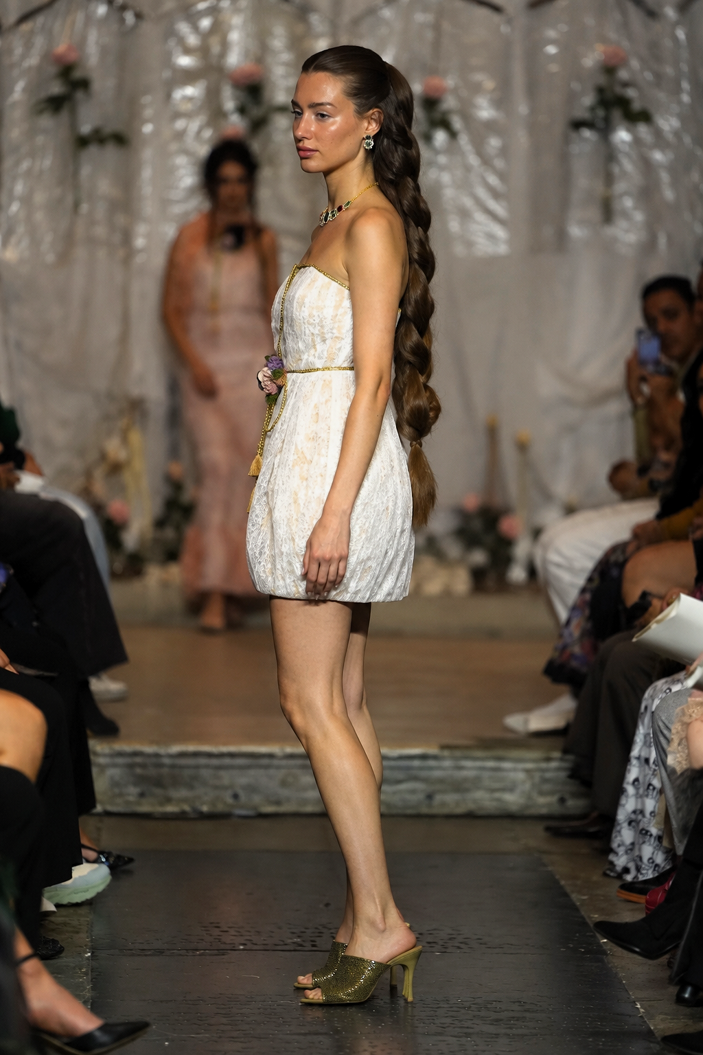A model walks the runway wearing a strapless white textured mini dress with a gold waist detail and olive green heeled mule sandals. The runway is flanked by seated spectators, with a draped white fabric backdrop in the background.