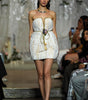 A model walks the runway wearing a strapless white textured mini dress with gold trim and a tassel tie at the waist, photographed during a fashion show with seated spectators in the background.